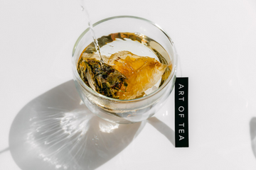 Hot water being poured into a glass of tea using an Art of Tea sachet bag.